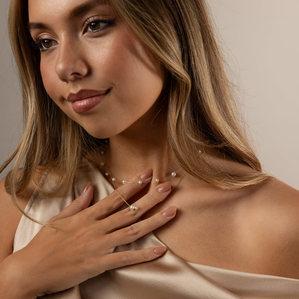 A woman with long hair, wearing a satin dress and the Floating Pearl Necklace, touches her chest and smiles softly.