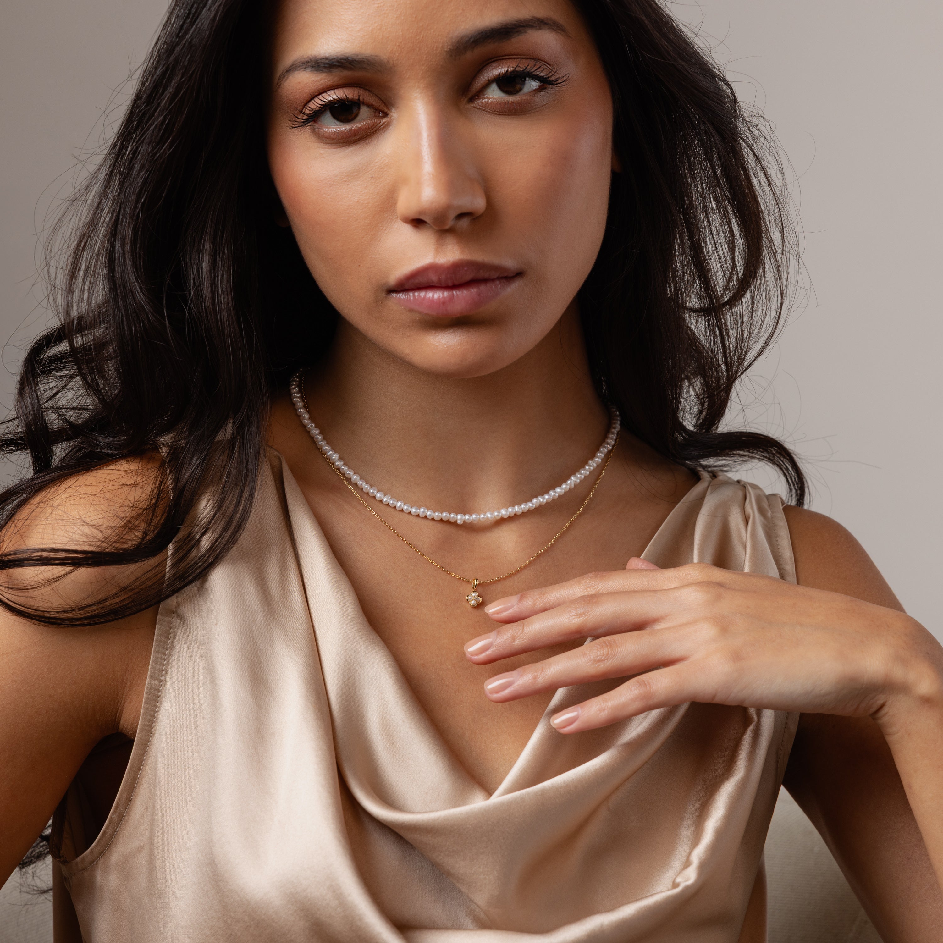 A woman in a beige satin top wears the Venus Pearl Necklace layered with gold chains, showcasing timeless jewelry as she looks at the camera.