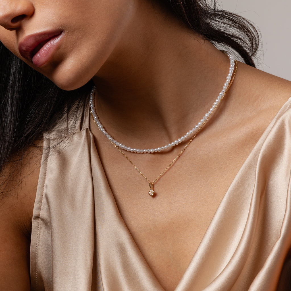 Woman in a silky beige top layers the Venus Pearl Necklace with a gold pendant, showcasing timeless jewelry elegance.