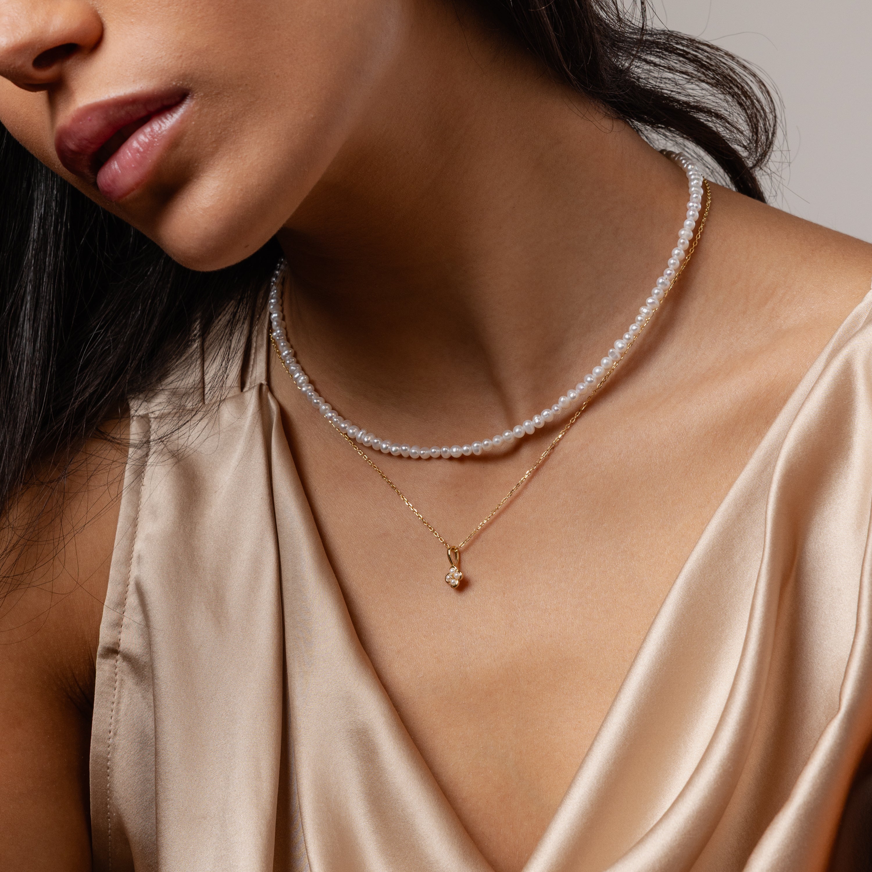 Woman in a silky beige top layers the Venus Pearl Necklace with a gold pendant, showcasing timeless jewelry elegance.
