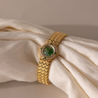 The Emerald Scalloped Timepiece features an emerald-green dial with diamond accents and a refined scallop-link bracelet, elegantly displayed on cream-colored fabric.