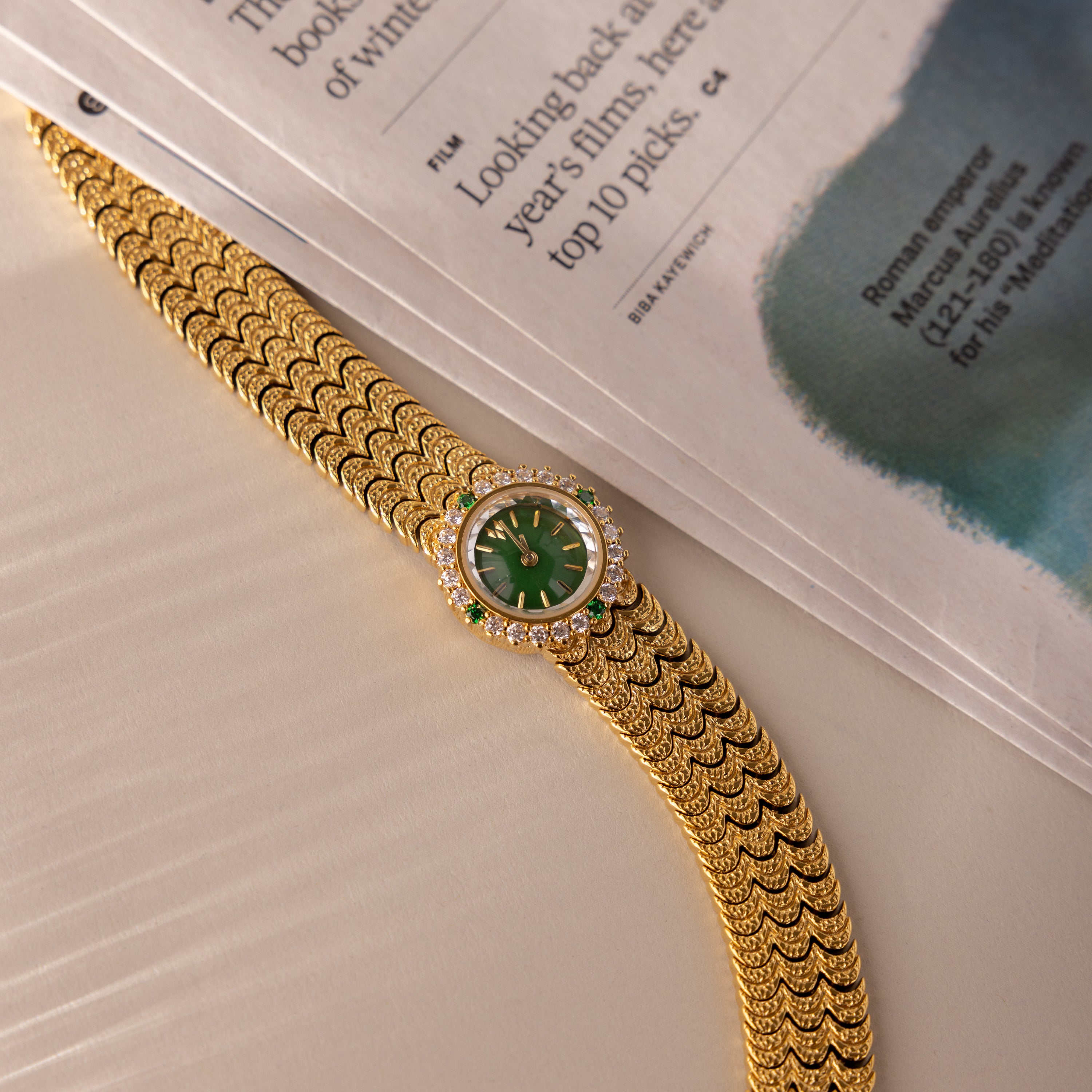 The Emerald Scalloped Timepiece, a gold wristwatch with an emerald-green dial and diamond accents, features a scallop-link bracelet and is displayed beside an open newspaper on a beige surface.