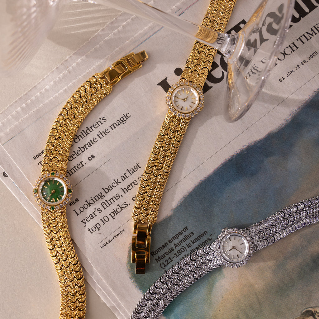 The Pave Scalloped Timepiece is displayed on a newspaper amid two other elegant wristwatches, with a wine glass tipped over it. It features a sparkling pavé bezel and a sophisticated scallop-link bracelet.