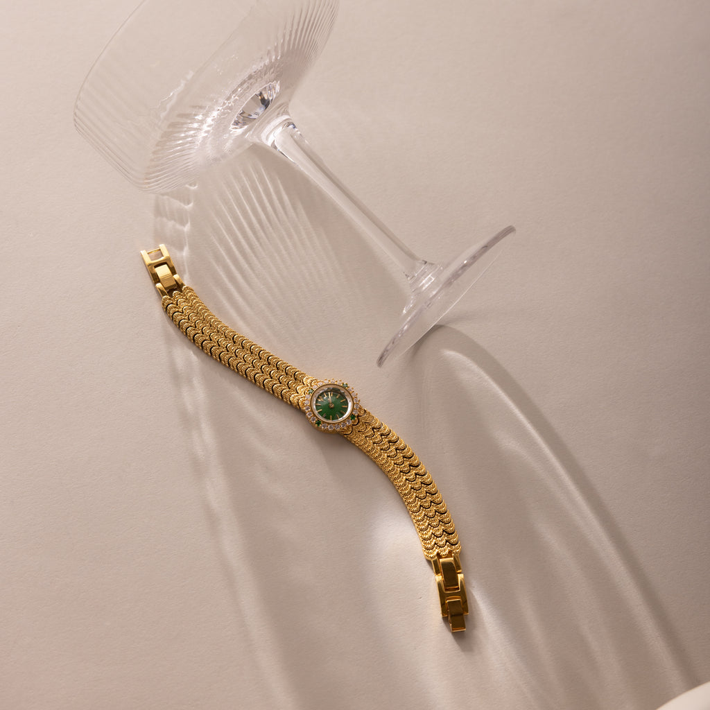 The Emerald Scalloped Timepiece, featuring a scallop-link bracelet and emerald-green dial, sits next to a tipped-over empty wine glass on a beige surface.