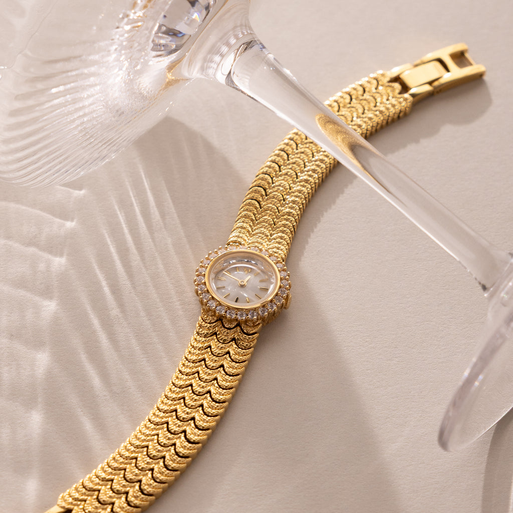 The Pave Scalloped Timepiece in 18K Gold, featuring a scallop-link bracelet and pavé bezel, rests beside clear wine glasses on a beige surface.