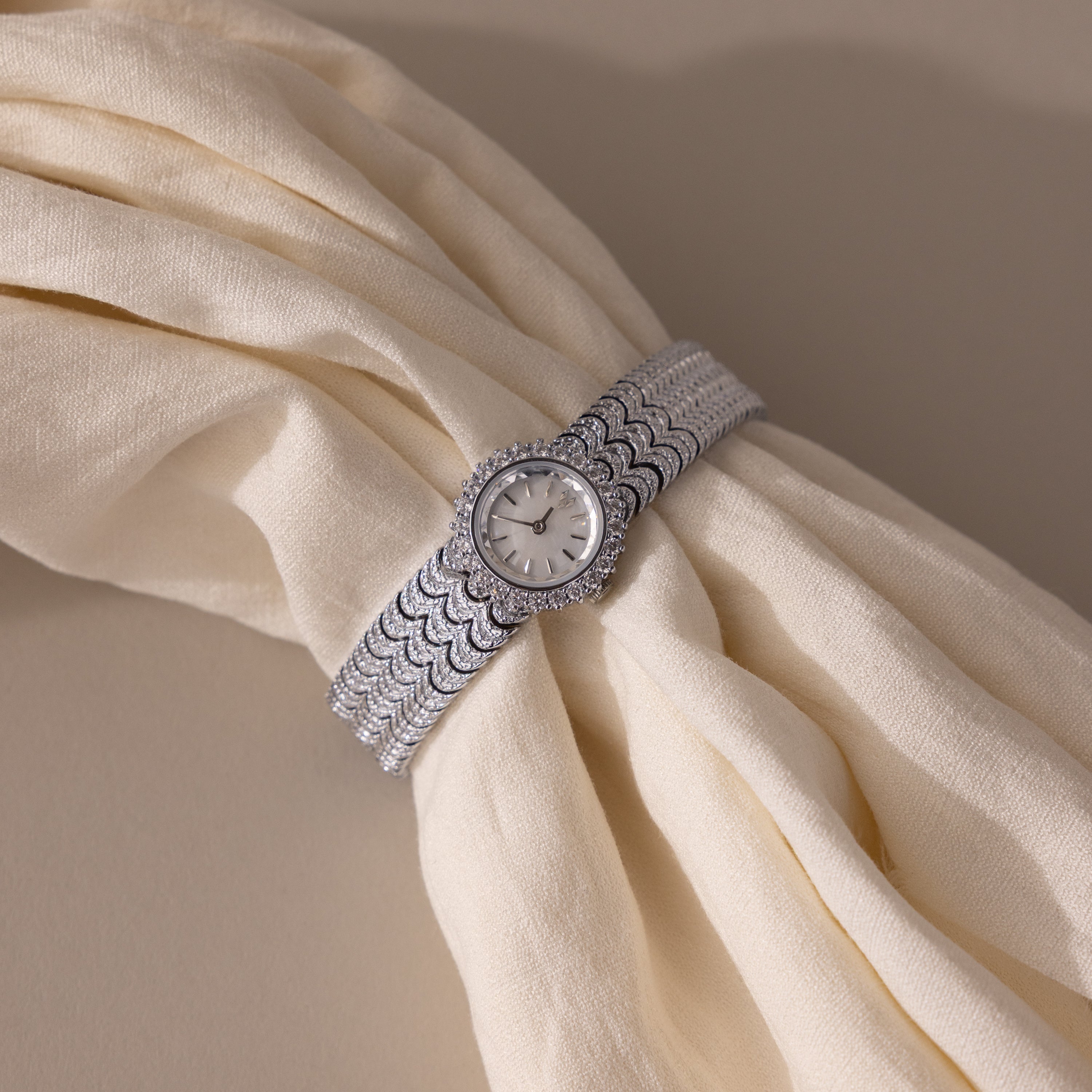 A Pave Scalloped Timepiece in White Gold with a pavé bezel is displayed on cream-colored, folded fabric.