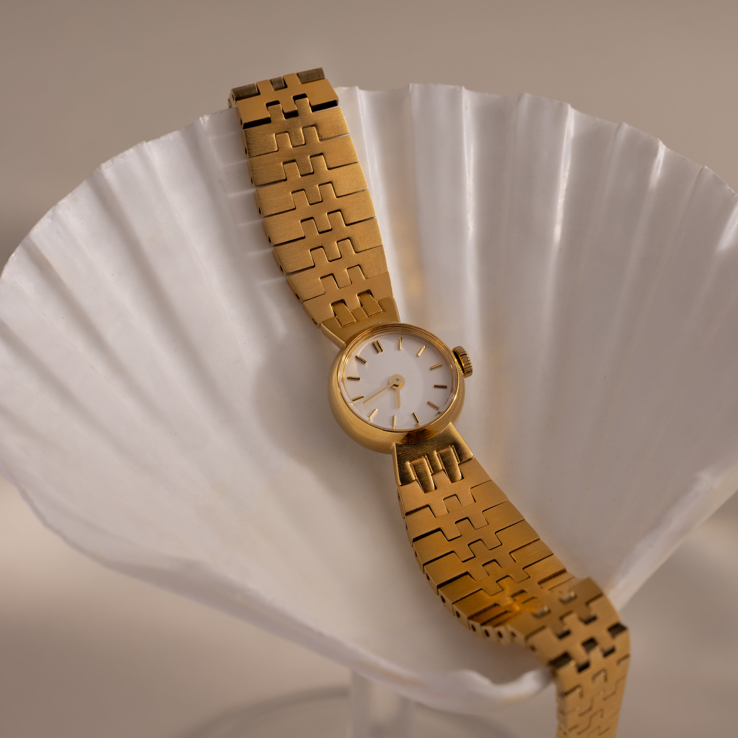 The Brick Link Timepiece features a sculptural gold case, white face, and bold brick-link bracelet, elegantly displayed in a white seashell for a modern, sophisticated look.