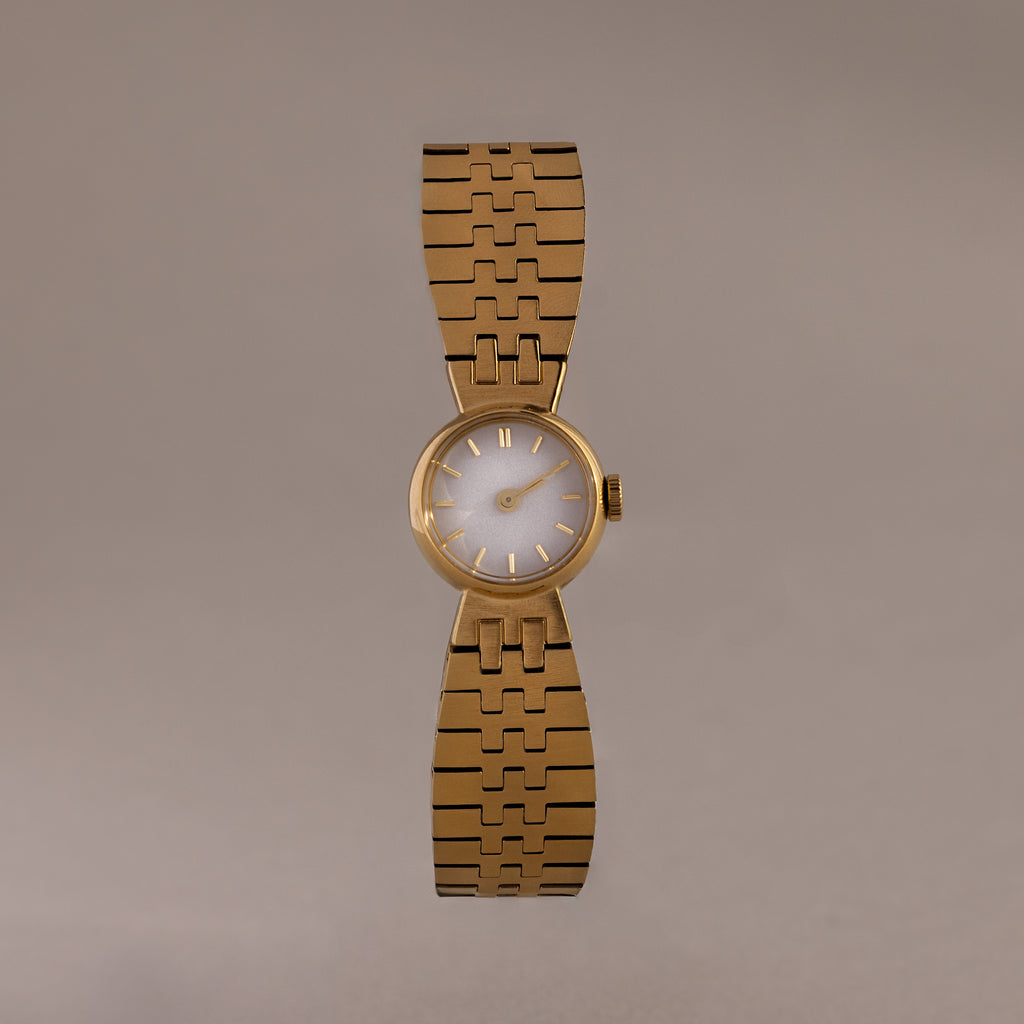 The Brick Link Timepiece is a gold sculptural watch with a round face and bold brick-link bracelet, showcased on a plain, light brown background.