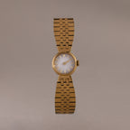 The Brick Link Timepiece is a gold sculptural watch with a round face and bold brick-link bracelet, showcased on a plain, light brown background.
