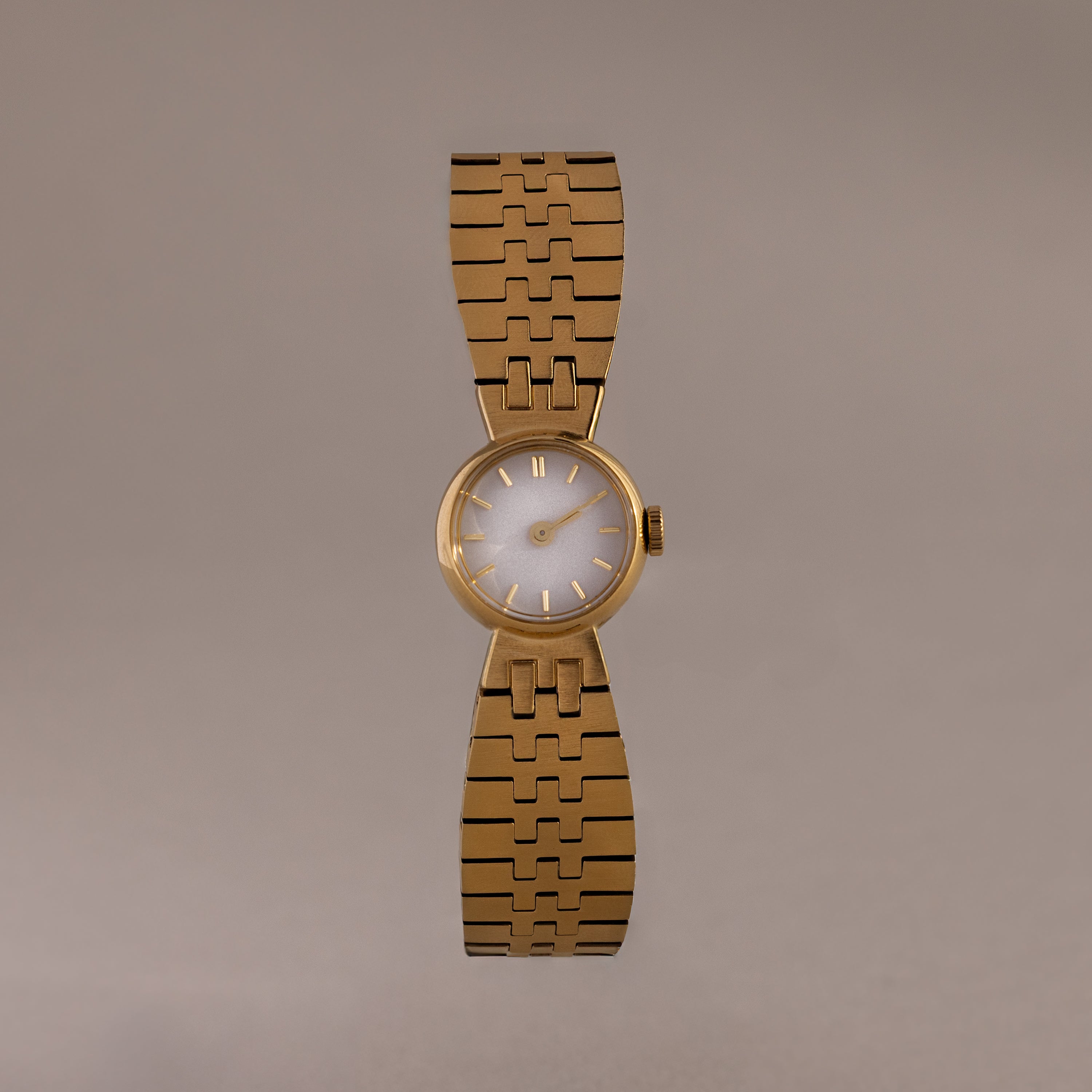 The Brick Link Timepiece is a gold sculptural watch with a round face and bold brick-link bracelet, showcased on a plain, light brown background.
