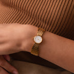 The Brick Link Timepiece, featuring a gold brick-link bracelet and white face, graces a person’s wrist against a brown ribbed top.