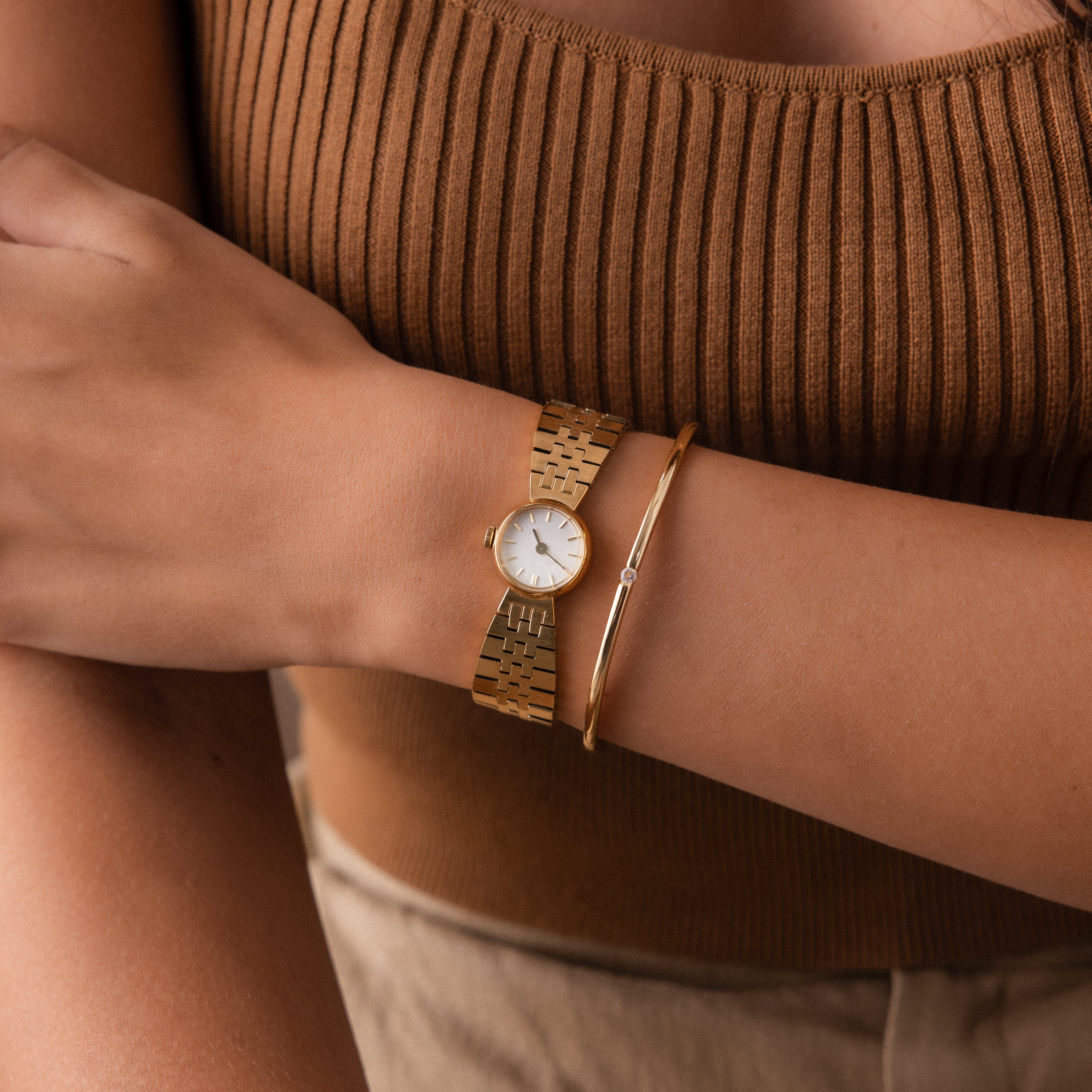 A person wearing the Brick Link Timepiece and a bold bracelet, paired with a brown ribbed tank top and beige pants.