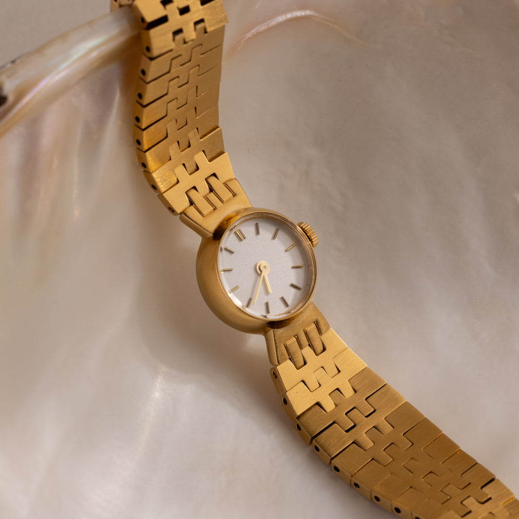 The Brick Link Timepiece, featuring a gold finish and white face, is elegantly displayed on a smooth, iridescent shell surface.