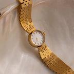 The Brick Link Timepiece, featuring a gold finish and white face, is elegantly displayed on a smooth, iridescent shell surface.