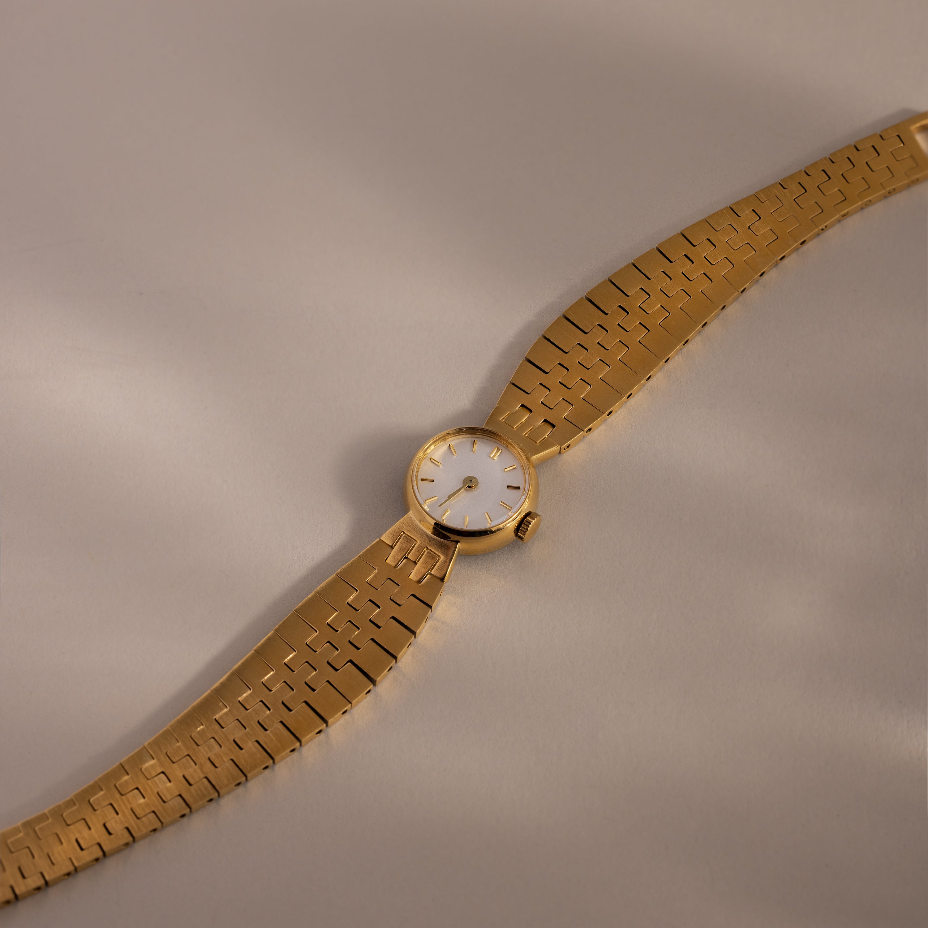The Brick Link Timepiece, featuring a white face and bold brick-link bracelet, is displayed diagonally on a light beige surface.