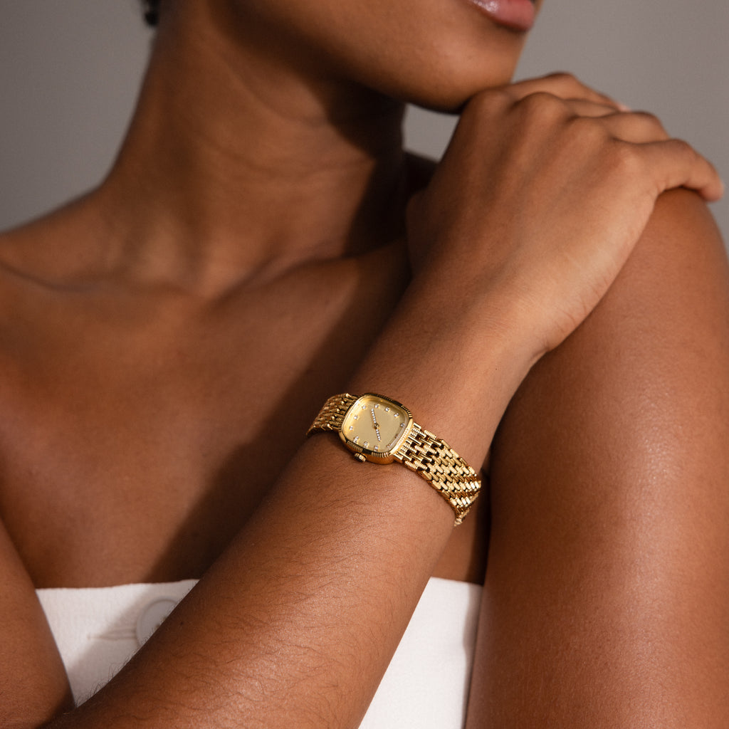A woman wearing the Pave Woven Timepiece with gold pavé detailing rests her arm across her chest, dressed in a white strapless top.