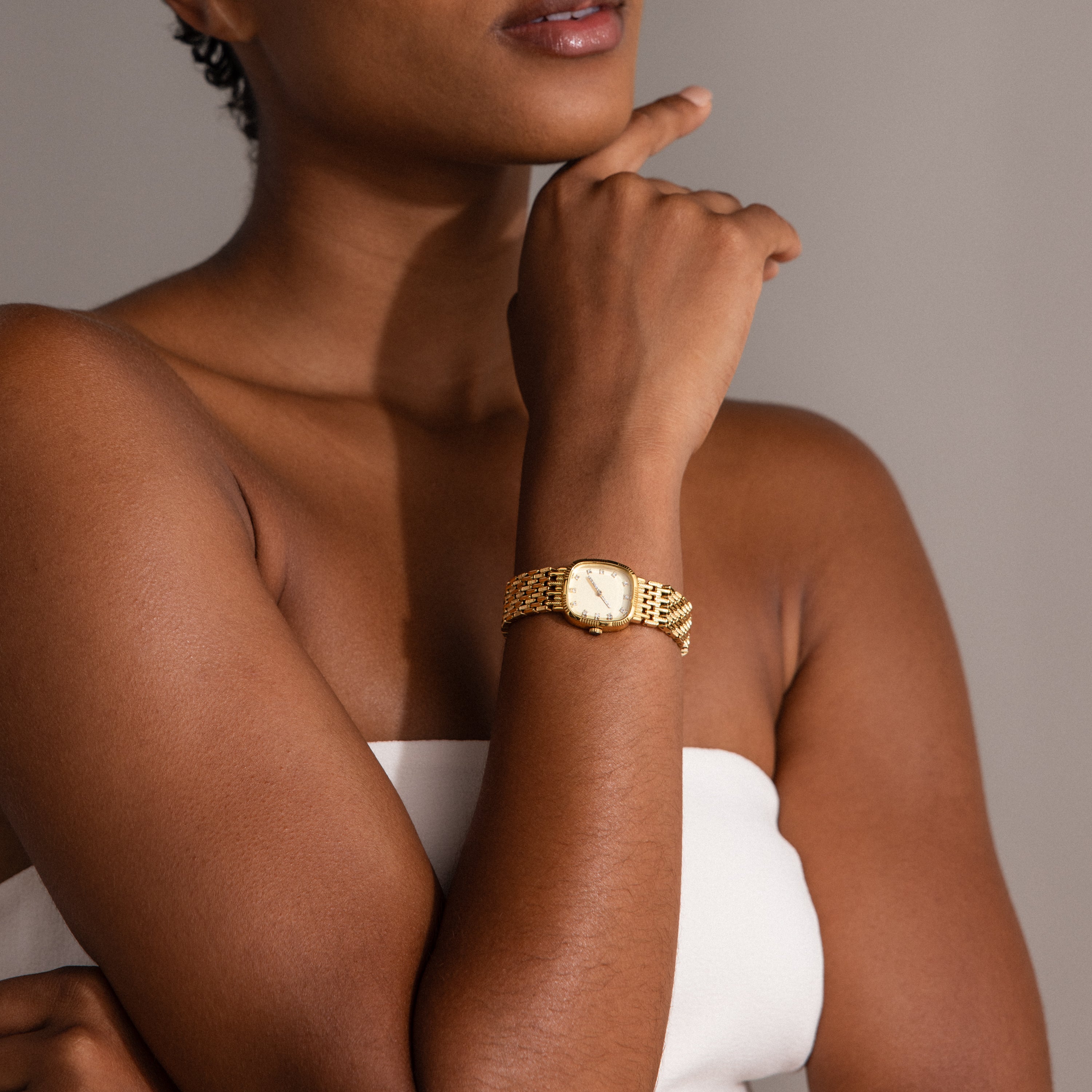 A woman in a white strapless top thoughtfully rests her chin on her hand, showcasing the Pave Woven Timepiece—a gold wristwatch with pavé detailing.