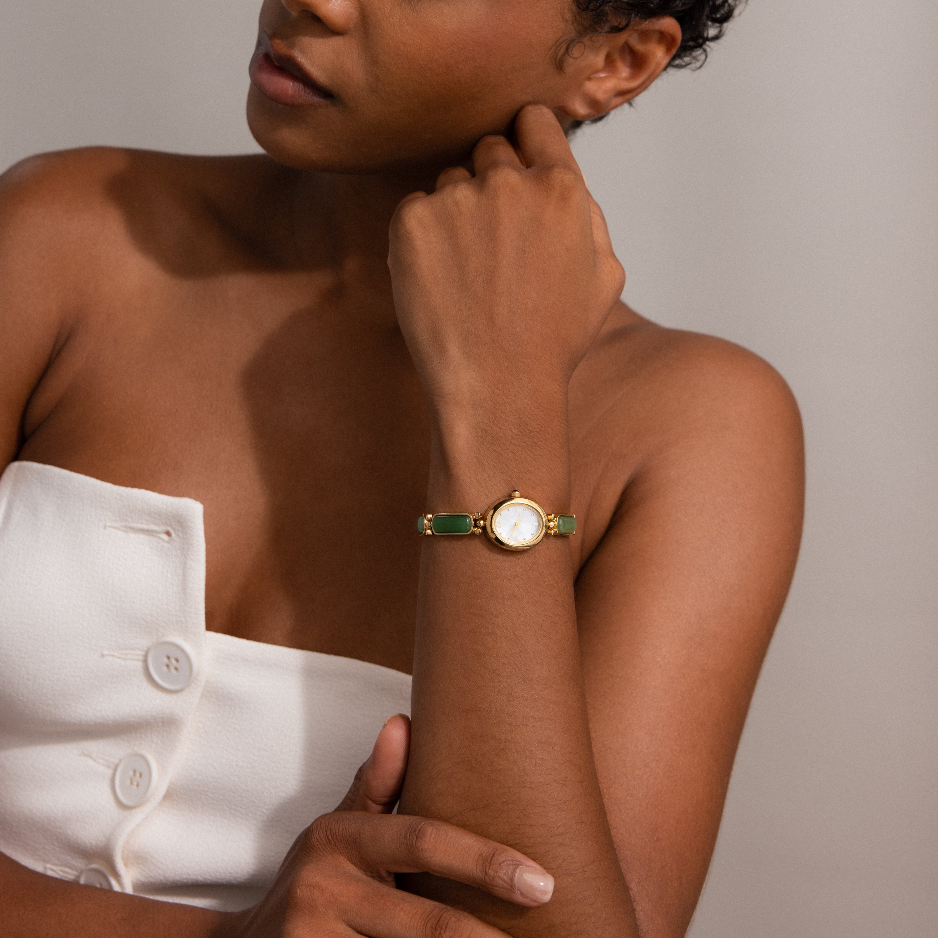 A woman in a white strapless top thoughtfully touches her neck while wearing the Jade Band Timepiece, a gold watch with an elegant jade band.