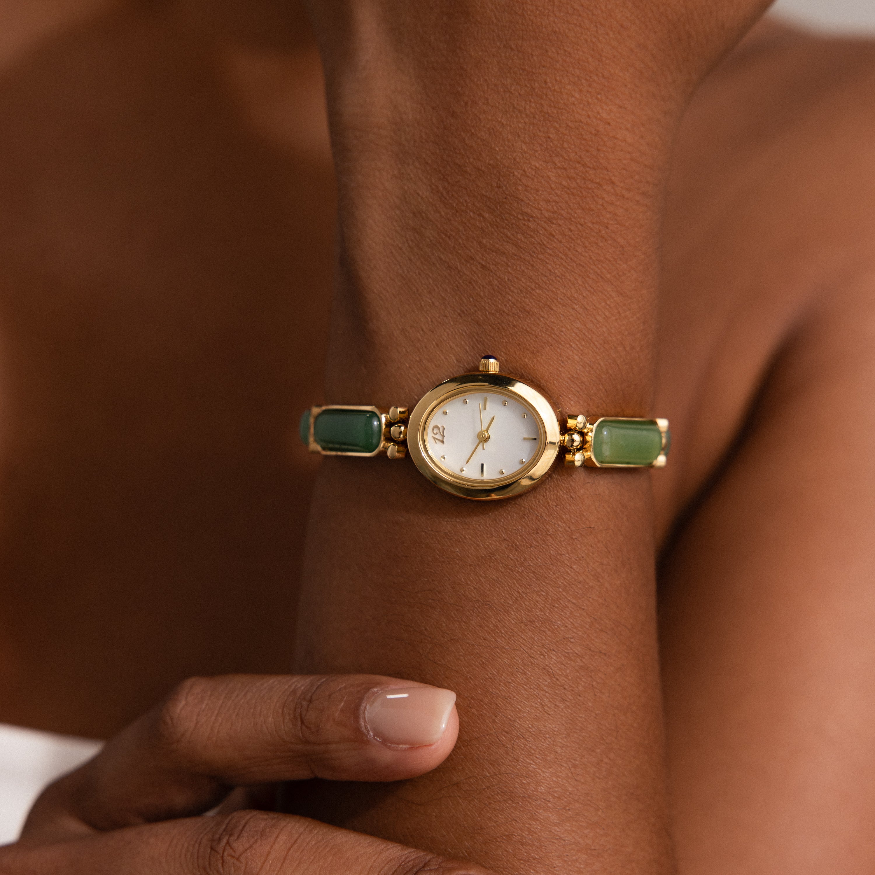 A person wears the Jade Band Timepiece, a gold wristwatch featuring a white minimalist oval dial and green jade band accents, while touching their arm.