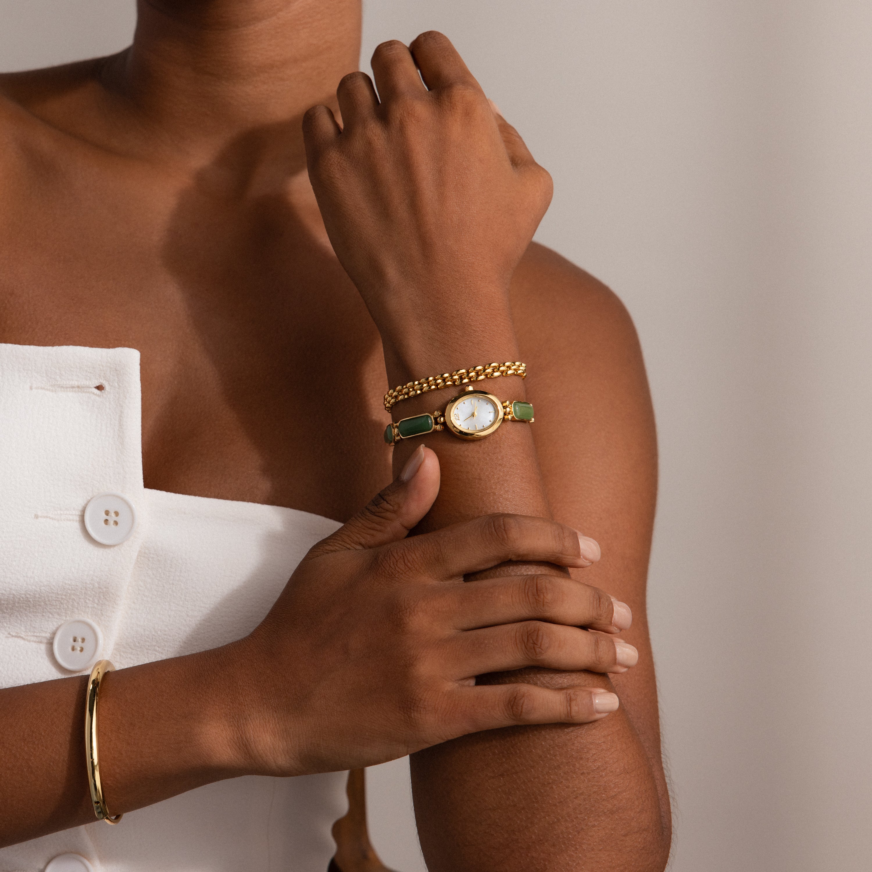 A woman wearing the Jade Band Timepiece, a gold watch with a minimalist oval dial, paired with a gold bracelet and a white buttoned dress, crosses her arms.