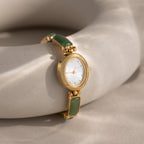 The Jade Band Timepiece features a gold wristwatch with a minimalist oval dial and green stone accents, elegantly displayed on a smooth beige surface. Its jade link bracelet adds refined style to this timeless design.