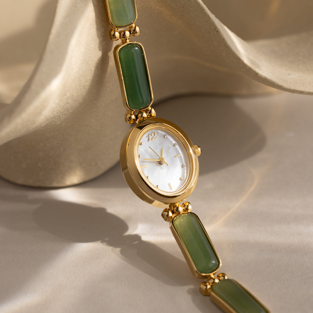 Jade Band Timepiece featuring a gold oval dial and green jade link bracelet, showcased on a light beige textured surface.