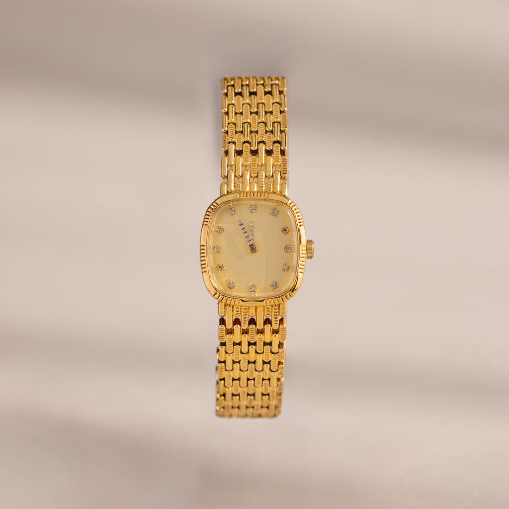 The Pave Woven Timepiece features a gold rectangular face, diamond hour markers, and pavé accents on a woven bracelet, elegantly displayed against a neutral background.