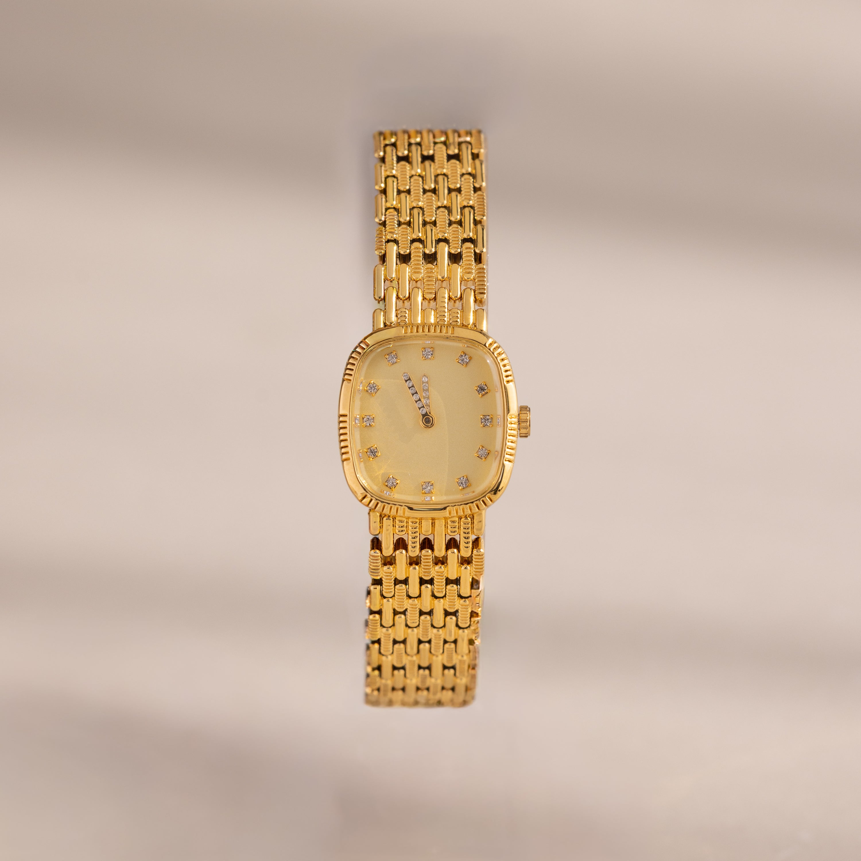 The Pave Woven Timepiece features a gold rectangular face, diamond hour markers, and pavé accents on a woven bracelet, elegantly displayed against a neutral background.