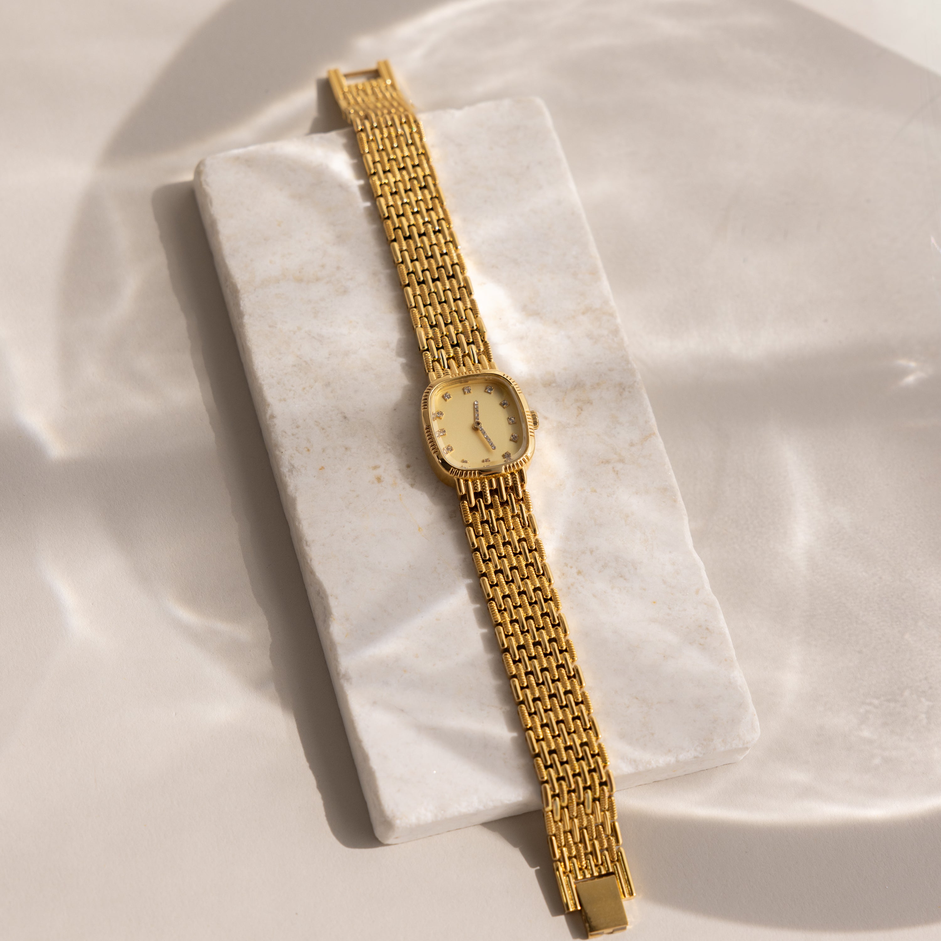 The Pave Woven Timepiece, featuring a gold linked band and pavé accents, is elegantly displayed on a rectangular white marble slab under soft lighting.