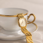 The Pave Woven Timepiece rests elegantly over the rim of a white teacup adorned with a gold handle and trim.