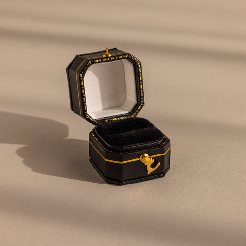 The Black Leather Ring Box, featuring gold accents and a velvet ring slot, sits open and empty on a beige surface under soft lighting.