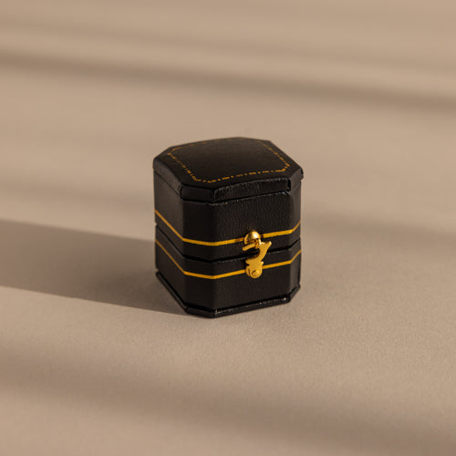 The Black Leather Ring Box is a small, black hexagonal jewelry box with yellow accents, a gold clasp, and a velvet ring slot, shown resting on a beige surface.