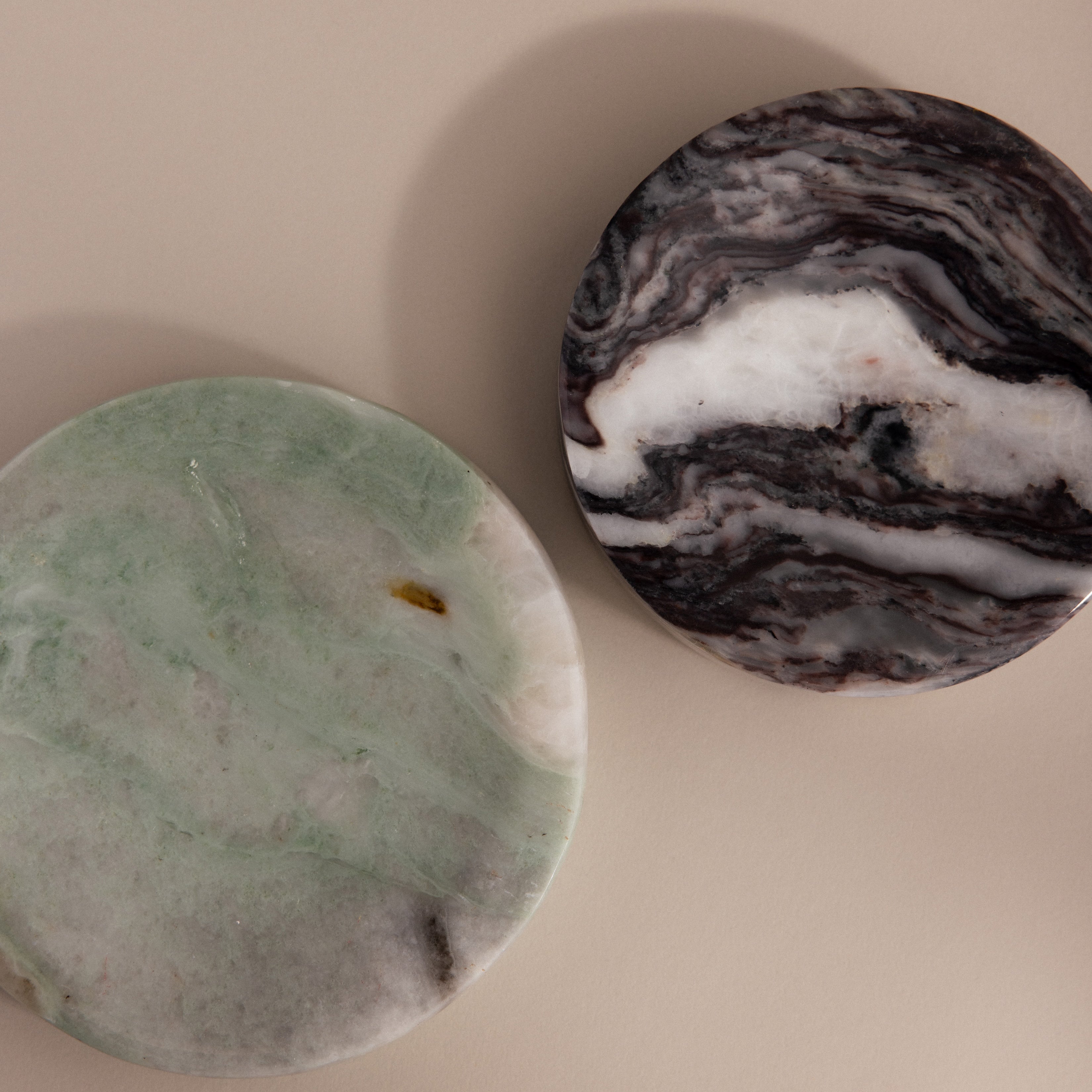 The Marble Jewelry Dish set includes two round marble coasters—one green and white, one black and white—on a beige surface, perfect for holding your favorite jewelry pieces in style.