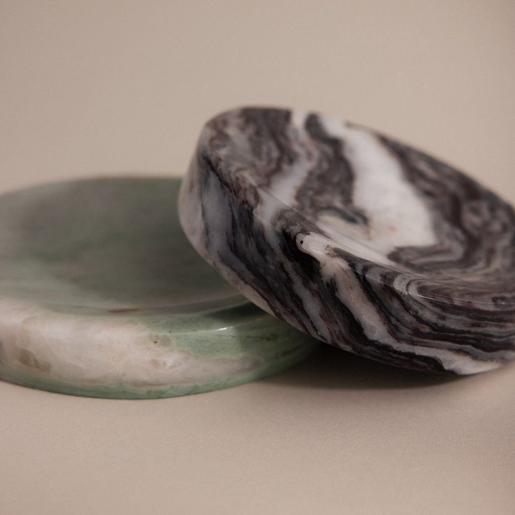 Two round, polished stone trays—one a greenish white jewelry holder, the other an Onyx Marble Jewelry Dish in black and white marbled pattern—rest elegantly on a beige background.