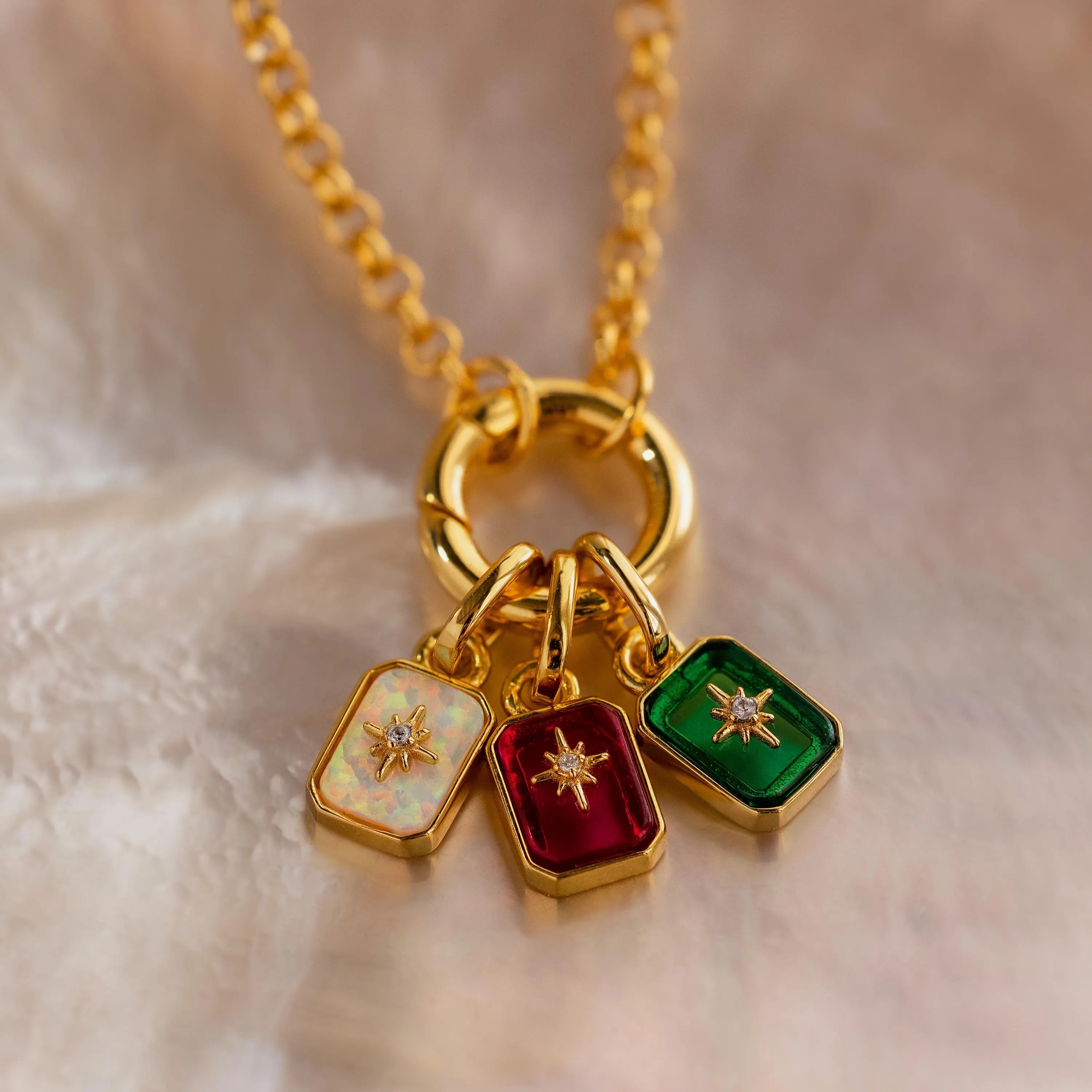 Up close of 3 north star birthstone charms in iridescent opal, red garnet, and green emerald, hooked on a gold rolo charm bracelet.