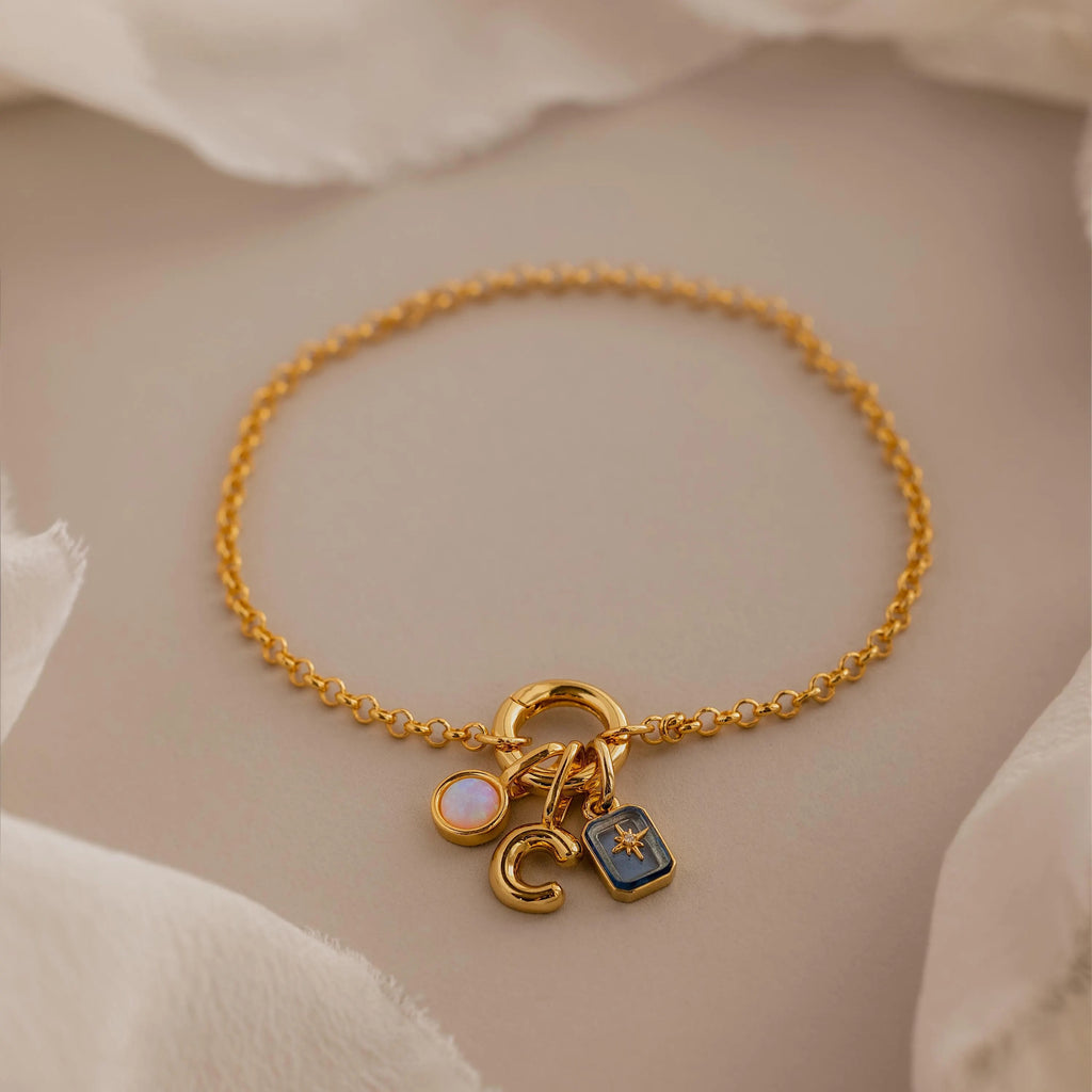 Dainty gold rolo charm bracelet with an opal signet charm, bubble letter "C" charm, and aquamarine north star charm displayed on a neutral background.