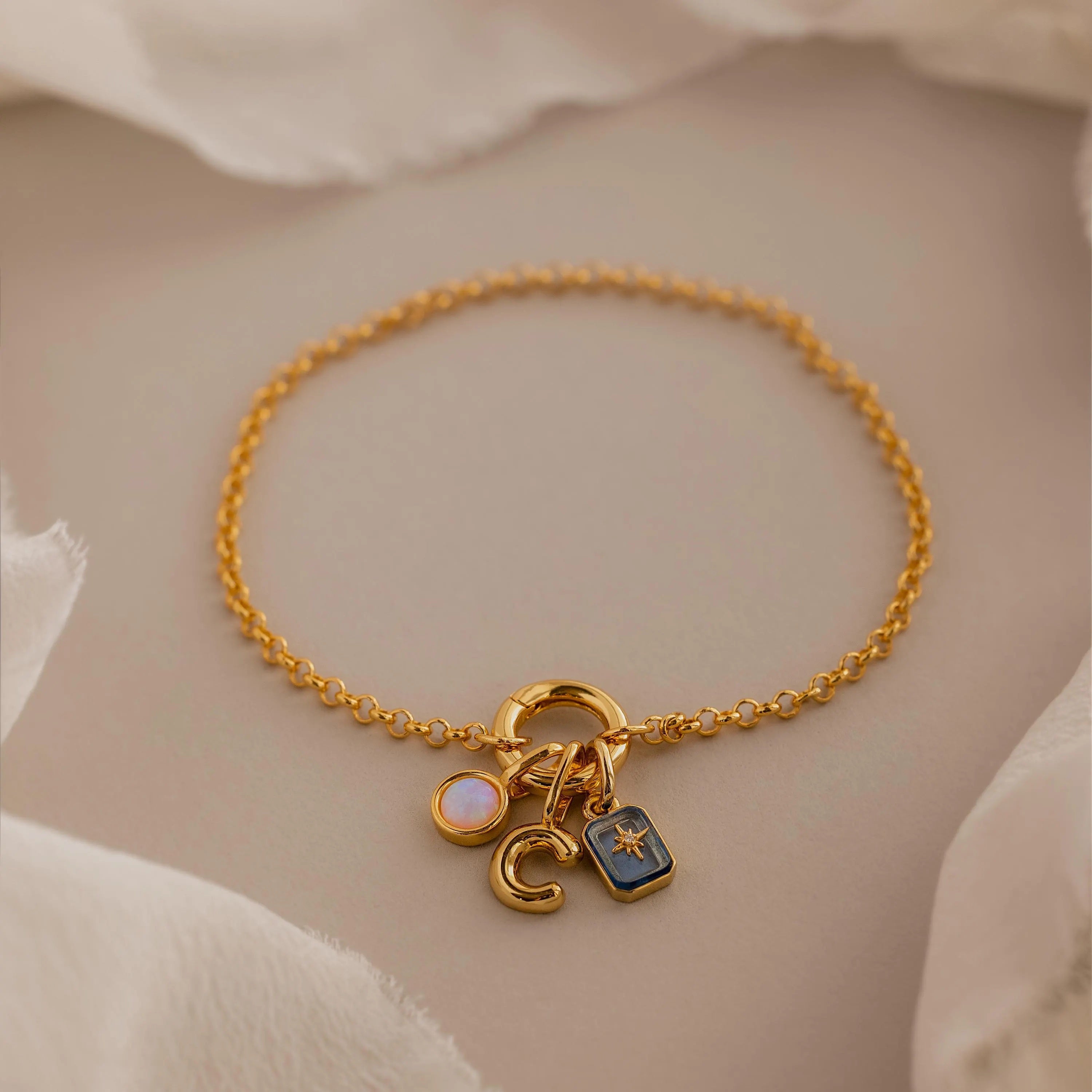 Dainty gold rolo charm bracelet with an opal signet charm, bubble letter "C" charm, and aquamarine north star charm displayed on a neutral background.