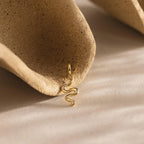 Single gold viper charm resting against the hollow curve of a textured, clay-toned ceramic prop.