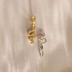 A close-up of two Pavé Viper Charms lying flat on a smooth, neutral-toned fabric. One charm is crafted in warm 18K gold and the other in cool sterling silver, both shaped like stylized serpents with flowing curves and micro pavé crystal details along the head and tail.