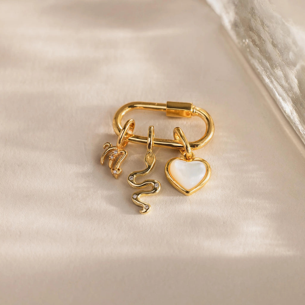Three gold charms—an evil eye charm, viper snake, and a glossy mother-of-pearl heart—hang from a gold carabiner clasp