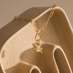 Gold paperclip chain necklace draped across a sculpted, wave-like ceramic prop. The Pavé Viper Charm is grouped with other symbolic pieces including an evil eye charm and an initial pearl charm, creating a balanced, elegant drop cluster.