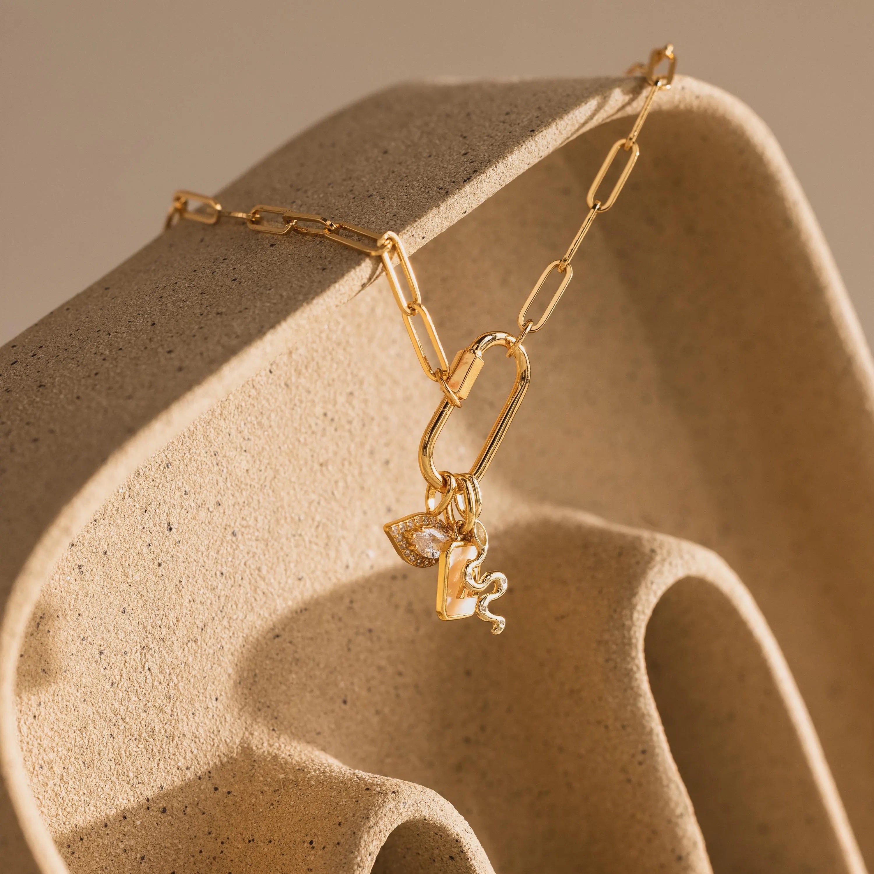 Gold paperclip chain necklace draped across a sculpted, wave-like ceramic prop. The Pavé Viper Charm is grouped with other symbolic pieces including an evil eye charm and an initial pearl charm, creating a balanced, elegant drop cluster.