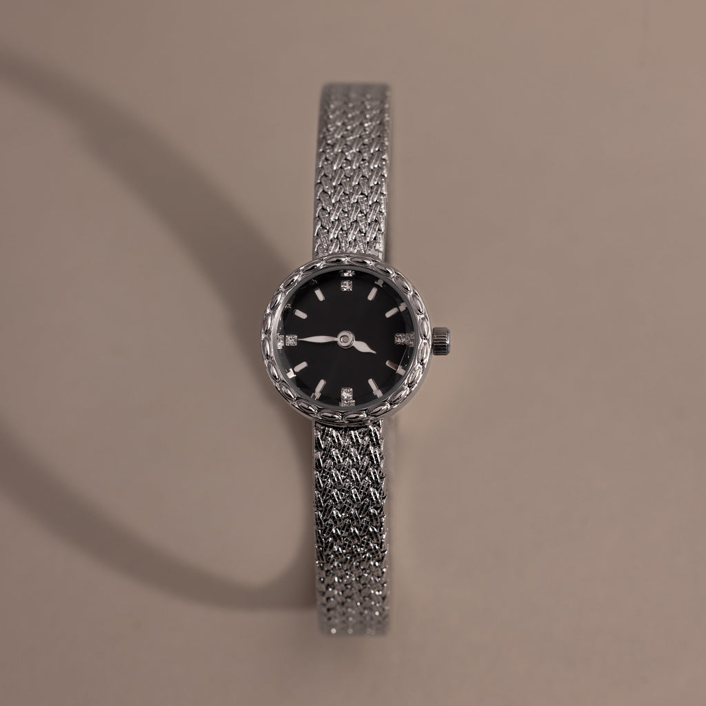 Silver braided timepiece with a black face centered on a neutral background.