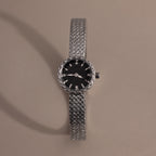 Silver braided timepiece with a black face centered on a neutral background.