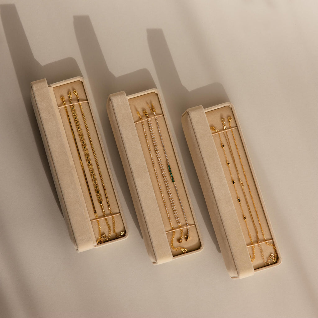 Three beige Bracelet Travel Cases hold delicate gold bracelets and necklaces, casting soft shadows on a light surface.
