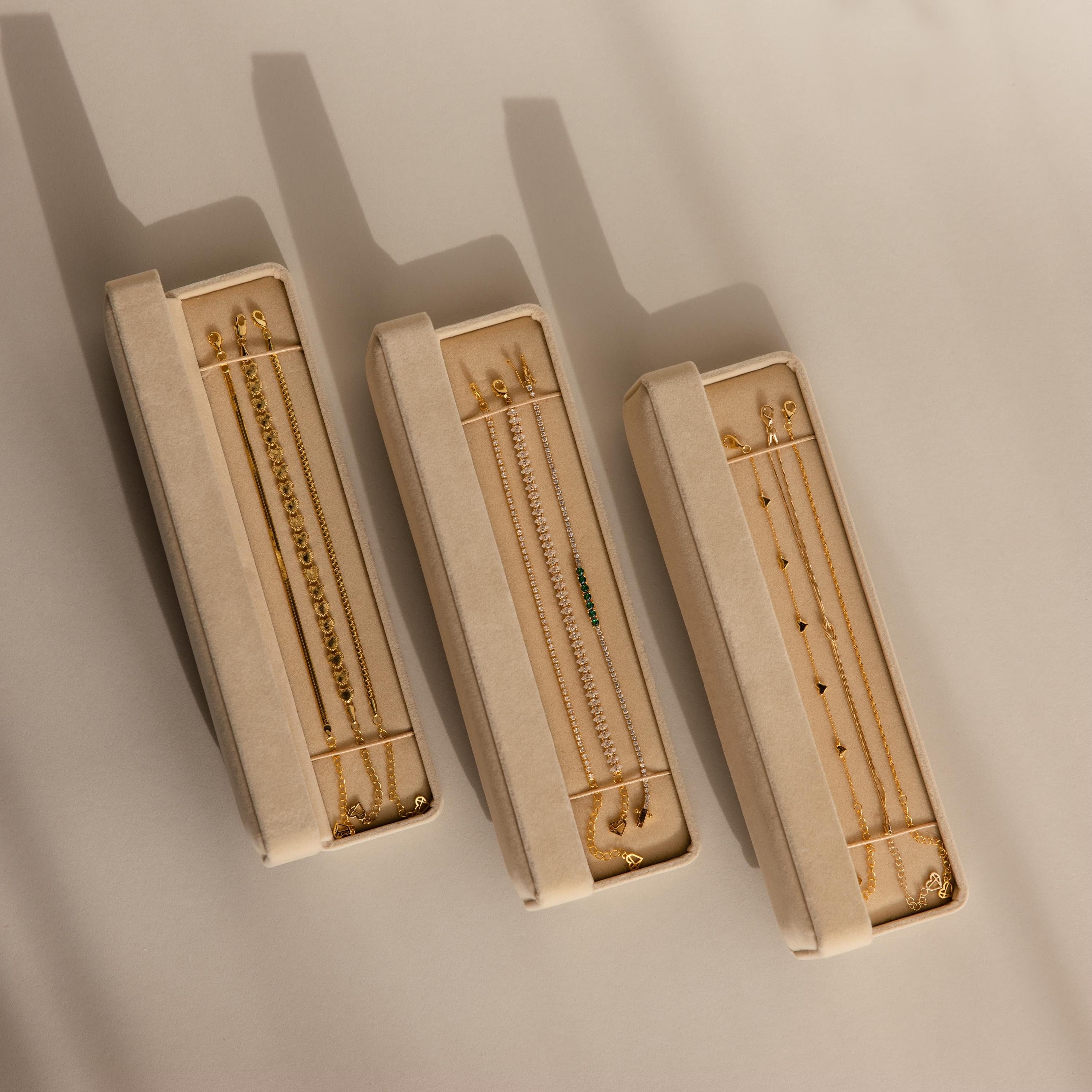 Three beige Bracelet Travel Cases hold delicate gold bracelets and necklaces, casting soft shadows on a light surface.