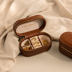 The Oval Leather Jewelry Box is a compact brown leather travel case for displaying gold rings and earrings on a beige fabric interior.