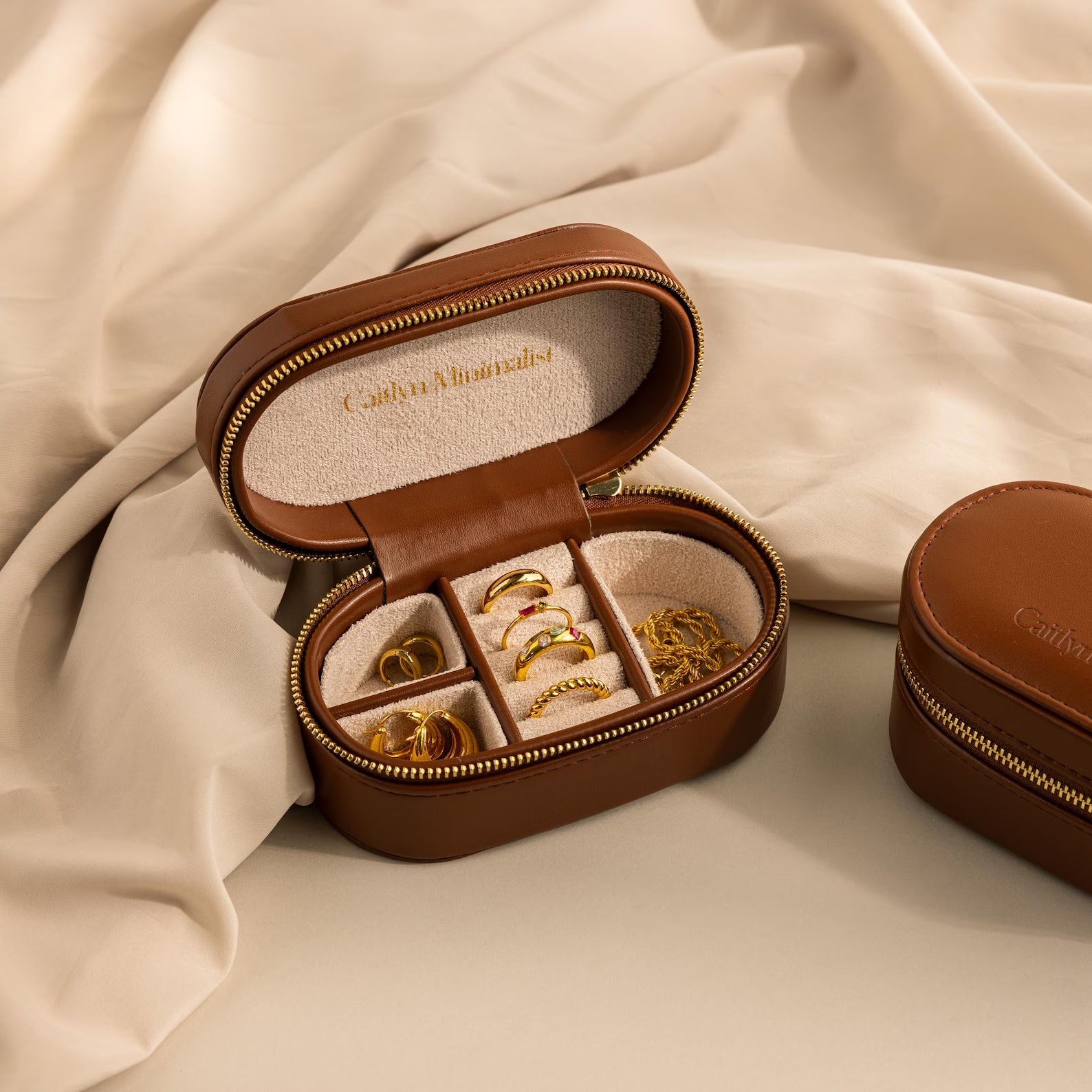 The Oval Leather Jewelry Box is a compact brown leather travel case for displaying gold rings and earrings on a beige fabric interior.