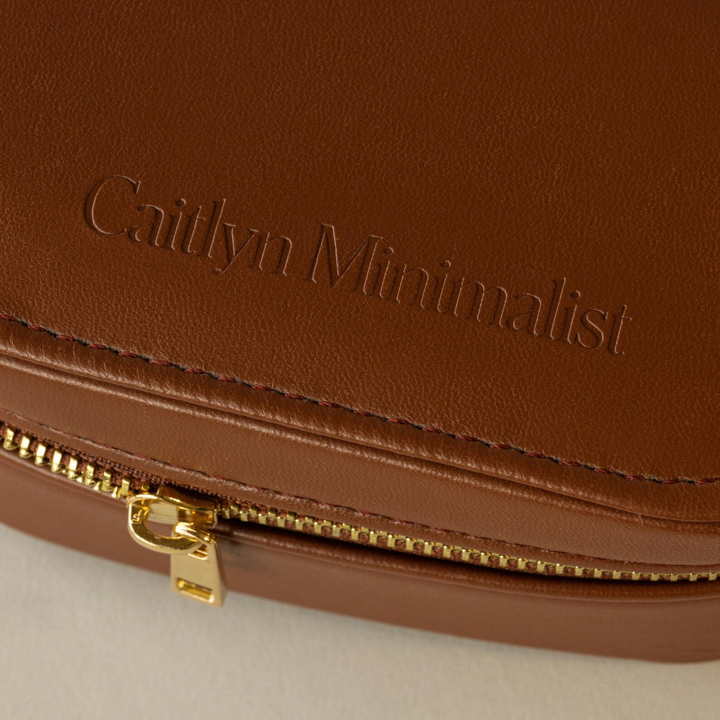Close-up of an Oval Leather Jewelry Box, a brown leather pouch embossed with Caitlyn Minimalist and a gold zipper, perfect as a compact travel jewelry organizer.