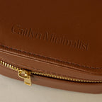Close-up of an Oval Leather Jewelry Box, a brown leather pouch embossed with Caitlyn Minimalist and a gold zipper, perfect as a compact travel jewelry organizer.